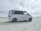A white van is parked on a sandy beach with overcast skies. The side window has text advertisements or promotional statements. The background features open space with another vehicle in the distance.