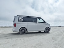 A white van is parked on a sandy beach with overcast skies. The side window has text advertisements or promotional statements. The background features open space with another vehicle in the distance.