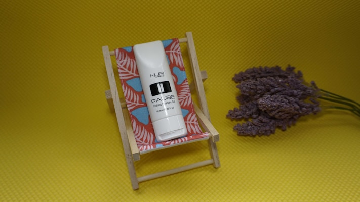 A small deck chair with a colorful red and blue patterned fabric holds a white tube labeled with the brand name and product details. Lavender flowers rest on the bright yellow background beside the chair.