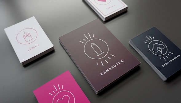 Several stacks of cards are laid out on a dark surface, each featuring different cover designs and labels. The cards include icons such as a cupcake with a candle, a heart, a condom, and a lightning cloud. The titles on the cards include 'Level 1,' 'Kamasutra,' and 'Gamechanger.' The cards have backgrounds in different colors such as white, black, and pink.