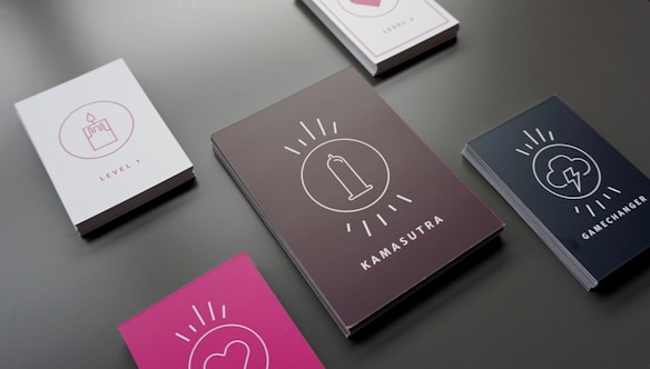 Several stacks of cards are laid out on a dark surface, each featuring different cover designs and labels. The cards include icons such as a cupcake with a candle, a heart, a condom, and a lightning cloud. The titles on the cards include 'Level 1,' 'Kamasutra,' and 'Gamechanger.' The cards have backgrounds in different colors such as white, black, and pink.