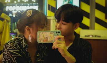 A couple is sitting closely together in a dimly lit setting. The girl has her hair tied up with a hair clip and is wearing large earrings and a patterned shirt. The boy is holding up a phone with a case decorated with various stickers. The background shows blurred lights and yellow-black striped patterns.