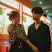 A young couple stands in front of brightly lit arcade claw machines. The woman, wearing glasses and a patterned shirt, has her hair styled up and is holding something. The man, wearing a striped shirt, is looking at her with a soft expression. The machines in the background are colorful, with signs displaying the word 'MEGABOX' and featuring cartoon characters.