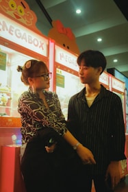 A young couple stands in front of brightly lit arcade claw machines. The woman, wearing glasses and a patterned shirt, has her hair styled up and is holding something. The man, wearing a striped shirt, is looking at her with a soft expression. The machines in the background are colorful, with signs displaying the word 'MEGABOX' and featuring cartoon characters.