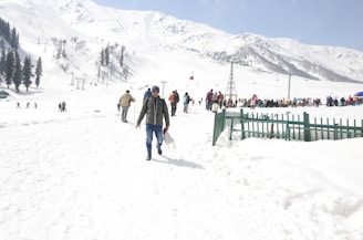 A snowy mountain landscape with numerous people engaged in winter activities such as skiing and walking. The scene includes ski lifts, snow-covered trees, and a distant mountain range. A green metal fence is visible in the snow, and some individuals are dressed in winter clothing, carrying equipment.