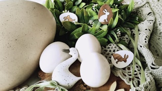 A collection of decorative Easter-themed items arranged on a soft, green crochet fabric. The setup includes three smooth, white eggs and a speckled duck figurine. Nestled among the eggs are wooden cut-outs shaped like rabbits and eggs, some with painted designs. Fresh green leaves and small white buds add to the natural and festive ambiance.