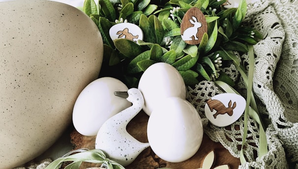 A collection of decorative Easter-themed items arranged on a soft, green crochet fabric. The setup includes three smooth, white eggs and a speckled duck figurine. Nestled among the eggs are wooden cut-outs shaped like rabbits and eggs, some with painted designs. Fresh green leaves and small white buds add to the natural and festive ambiance.