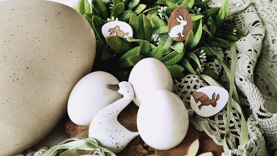 A collection of decorative Easter-themed items arranged on a soft, green crochet fabric. The setup includes three smooth, white eggs and a speckled duck figurine. Nestled among the eggs are wooden cut-outs shaped like rabbits and eggs, some with painted designs. Fresh green leaves and small white buds add to the natural and festive ambiance.