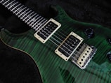A close-up view of an electric guitar with a green tiger stripe finish. The guitar features dual humbucker pickups, a rosewood fretboard with distinctive inlays, and a tremolo bridge. The control knobs are visible and the strings run across the body, showing chrome and ivory hardware against the dark background.