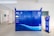 A blue package locker system is positioned in an indoor space. The locker has several compartments and a digital screen for interaction. There is a sign on the locker with various service instructions. A small stand with safety messages is located next to it. The setting is clean and minimalistic, with light-colored walls and floor tiles.