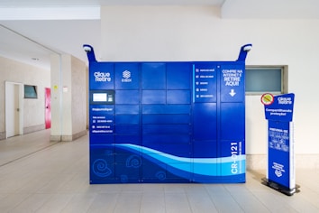 A blue package locker system is positioned in an indoor space. The locker has several compartments and a digital screen for interaction. There is a sign on the locker with various service instructions. A small stand with safety messages is located next to it. The setting is clean and minimalistic, with light-colored walls and floor tiles.