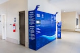 A bright interior space with a modern blue parcel locker system against a light-colored wall. The locker has detailed instructions in Portuguese and is accompanied by a small, informative stand on the right. On the left side, a red fire extinguisher is mounted on the wall near a men's restroom sign.