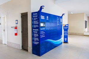 A bright interior space with a modern blue parcel locker system against a light-colored wall. The locker has detailed instructions in Portuguese and is accompanied by a small, informative stand on the right. On the left side, a red fire extinguisher is mounted on the wall near a men's restroom sign.