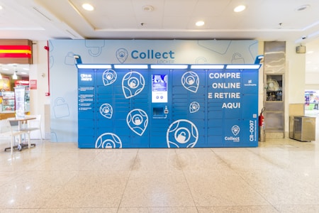 A blue parcel locker is installed in a shopping mall. The front of the locker features various compartments with a central touchscreen display. The logo and some text are printed in white. The area is well-lit with a tiled floor, and to the left, there is a small caf&eacute; table and chairs.