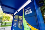 A brightly colored locker station with a blue canopy and walls marked with text 'E-BOX Clique Retire' and hashtags such as '#sejaumclicquer'. The locker is designed for parcel collection and is situated outdoors with greenery and buildings visible in the background.