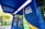 A brightly colored locker station with a blue canopy and walls marked with text 'E-BOX Clique Retire' and hashtags such as '#sejaumclicquer'. The locker is designed for parcel collection and is situated outdoors with greenery and buildings visible in the background.