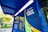A brightly colored locker station with a blue canopy and walls marked with text 'E-BOX Clique Retire' and hashtags such as '#sejaumclicquer'. The locker is designed for parcel collection and is situated outdoors with greenery and buildings visible in the background.