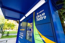 A brightly colored locker station with a blue canopy and walls marked with text 'E-BOX Clique Retire' and hashtags such as '#sejaumclicquer'. The locker is designed for parcel collection and is situated outdoors with greenery and buildings visible in the background.
