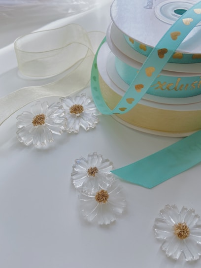 A collection of decorative ribbons in pastel shades is placed on a white surface. One ribbon is turquoise with golden heart patterns, while another is a delicate cream color. There are also translucent floral ornaments with gold centers scattered around the ribbons.