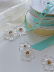 A collection of decorative ribbons in pastel shades is placed on a white surface. One ribbon is turquoise with golden heart patterns, while another is a delicate cream color. There are also translucent floral ornaments with gold centers scattered around the ribbons.