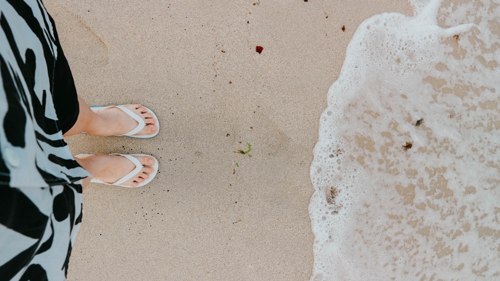 Person in braunen Flip-Flops steht tagsüber am weißen Sandstrand