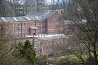 A large, historic brick building with numerous windows, surrounded by leafless trees and greenery. The structure has multiple levels and a roof with skylights, situated in a wooded rural setting.