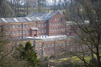 A large, historic brick building with numerous windows, surrounded by leafless trees and greenery. The structure has multiple levels and a roof with skylights, situated in a wooded rural setting.