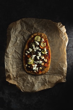 An artisanal flatbread garnished with crumbled white cheese, roasted vegetables like zucchini and possibly squash, is placed on a piece of crumpled brown parchment paper.