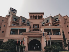 A large, multi-story building with architectural features resembling a hotel or resort. The structure showcases numerous balconies with decorative railings and arches, and is adorned with intricate tile work. Flags from different countries are positioned in front of the entrance, enhancing the international ambiance. Small shrubs and trees are placed around the base, adding greenery to the scene.