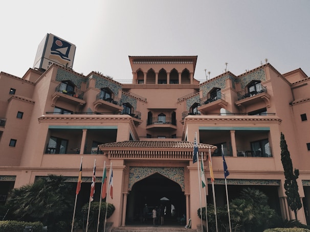 A large, multi-story building with architectural features resembling a hotel or resort. The structure showcases numerous balconies with decorative railings and arches, and is adorned with intricate tile work. Flags from different countries are positioned in front of the entrance, enhancing the international ambiance. Small shrubs and trees are placed around the base, adding greenery to the scene.