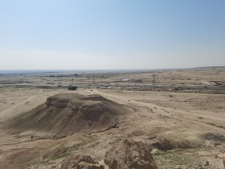 A vast desert landscape with a large sandy hill in the foreground. In the distance, power lines and towers are visible, with a road running parallel to them. Sparse vegetation appears scattered across the arid land under a clear blue sky.