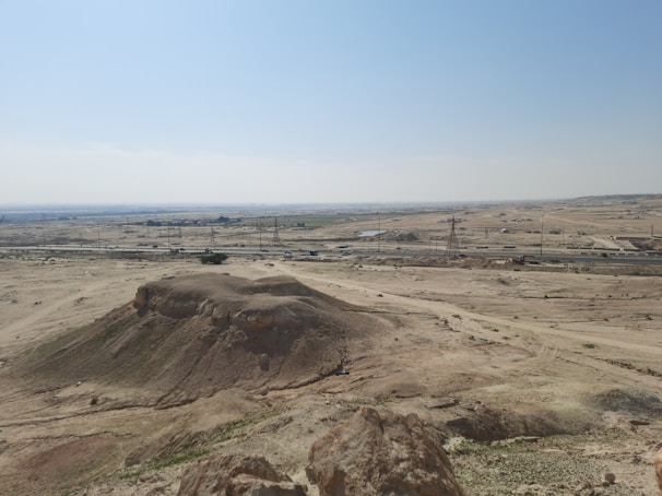 A vast desert landscape with a large sandy hill in the foreground. In the distance, power lines and towers are visible, with a road running parallel to them. Sparse vegetation appears scattered across the arid land under a clear blue sky.