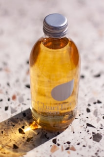 A clear glass bottle filled with a golden liquid, topped with a silver cap. The label shows text and a drop logo. It rests on a terrazzo surface, casting a long shadow. Light penetrates the liquid, creating a warm glow.