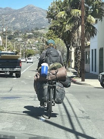 A cyclist carrying a large amount of baggage rides down a suburban street. The cyclist has a backpack and several bags secured to the bike, with a sleeping bag and a blue water container visible. The scene is set against a backdrop of mountains and palm trees lining the street, with other vehicles and buildings on either side.