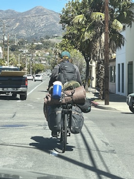 A cyclist carrying a large amount of baggage rides down a suburban street. The cyclist has a backpack and several bags secured to the bike, with a sleeping bag and a blue water container visible. The scene is set against a backdrop of mountains and palm trees lining the street, with other vehicles and buildings on either side.