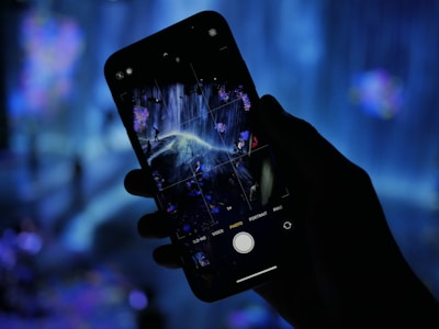 A hand is holding a smartphone capturing a digital art installation with flowing lights and abstract forms. The background is filled with a soft blue glow, contributing to the surreal ambience.