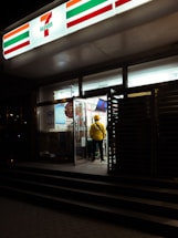 A person wearing a yellow jacket is standing near the entrance of a brightly lit 7-Eleven store at night. The glass doors are open, and various products can be seen inside.