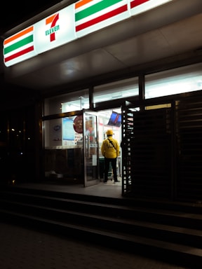 A person wearing a yellow jacket is standing near the entrance of a brightly lit 7-Eleven store at night. The glass doors are open, and various products can be seen inside.