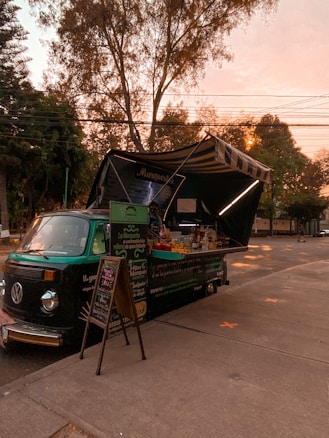 A vintage green food truck parked on the side of the road is serving food. The truck has open panels displaying various food items and a menu written on a blackboard. Trees with dense foliage surround the area, and the sun is setting in the background, casting a warm glow over the scene.