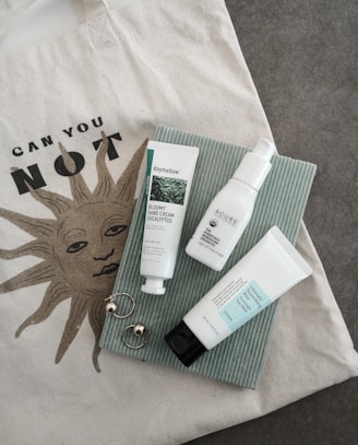 A soft, neutral-toned flat lay of a minimalist tote bag, sunglasses, and a delicate gold necklace on a white linen background.