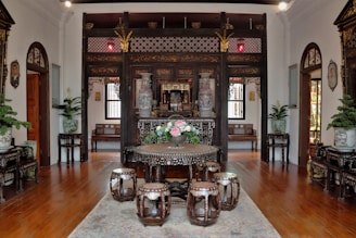 An intricately decorated room featuring dark wooden furniture with elaborate carvings and mother-of-pearl inlays. The centerpiece is a round table adorned with a floral arrangement surrounded by matching stools. Large vases with ornate designs and lush potted plants are positioned around the room, adding to the elegant Oriental aesthetic. The floor is wooden, accented by a soft rug beneath the table set. Traditional wall art and lanterns enhance the cultural ambiance.