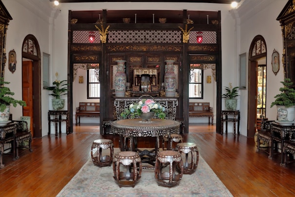An intricately decorated room featuring dark wooden furniture with elaborate carvings and mother-of-pearl inlays. The centerpiece is a round table adorned with a floral arrangement surrounded by matching stools. Large vases with ornate designs and lush potted plants are positioned around the room, adding to the elegant Oriental aesthetic. The floor is wooden, accented by a soft rug beneath the table set. Traditional wall art and lanterns enhance the cultural ambiance.
