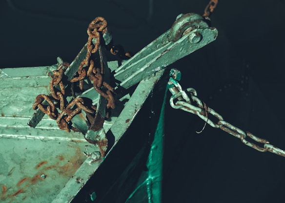 A close-up view of a rusty chain attached to the weathered metallic hull of a boat. The metal surface has a turquoise-green color with visible signs of rust and oxidation. The chain links are thick, prominently corroded, and connected to a metal fitting.
