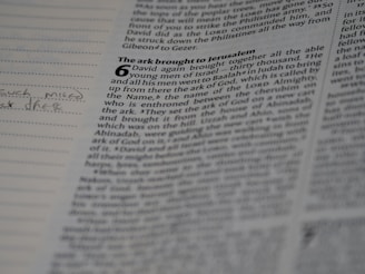 A close-up view of an open book with visible text. The page appears to be from the Bible, particularly featuring a passage with a large number '6' and a chapter title 'The ark brought to Jerusalem'. There is handwritten text in the left margin of the page.