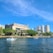 The image features a waterfront view with several boats on the water. In the background, a prominent building labeled 'Straz Center'. There are other tall buildings and skyscrapers as part of a city skyline. Lush green trees line the shore, contributing to an overall vibrant and sunny scene.