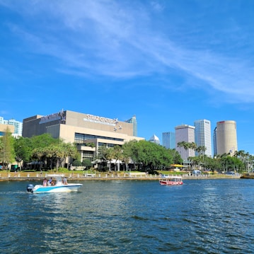The image features a waterfront view with several boats on the water. In the background, a prominent building labeled 'Straz Center'. There are other tall buildings and skyscrapers as part of a city skyline. Lush green trees line the shore, contributing to an overall vibrant and sunny scene.