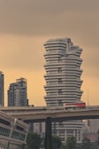 A large, uniquely designed white building with a stacked, angular structure stands prominently against a muted sky. In the foreground, a bridge carries a red double-decker bus with 'SBS Transit' written on it. Surrounding the main building are other modern skyscrapers, adding to the urban landscape. Lush green trees are visible at the bottom, juxtaposing the otherwise concrete environment.