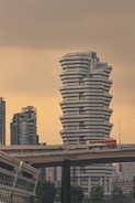 A large, uniquely designed white building with a stacked, angular structure stands prominently against a muted sky. In the foreground, a bridge carries a red double-decker bus with 'SBS Transit' written on it. Surrounding the main building are other modern skyscrapers, adding to the urban landscape. Lush green trees are visible at the bottom, juxtaposing the otherwise concrete environment.