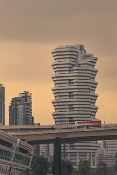 A large, uniquely designed white building with a stacked, angular structure stands prominently against a muted sky. In the foreground, a bridge carries a red double-decker bus with 'SBS Transit' written on it. Surrounding the main building are other modern skyscrapers, adding to the urban landscape. Lush green trees are visible at the bottom, juxtaposing the otherwise concrete environment.