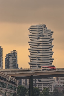 A large, uniquely designed white building with a stacked, angular structure stands prominently against a muted sky. In the foreground, a bridge carries a red double-decker bus with 'SBS Transit' written on it. Surrounding the main building are other modern skyscrapers, adding to the urban landscape. Lush green trees are visible at the bottom, juxtaposing the otherwise concrete environment.
